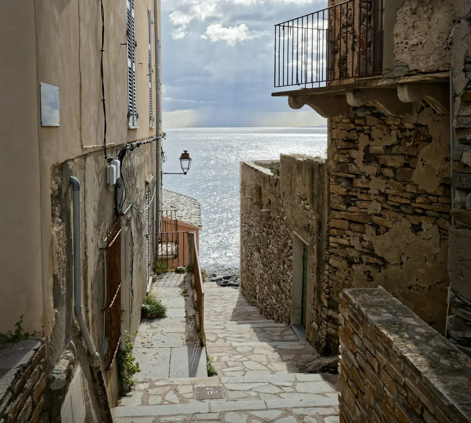 A stone lane between two old houses, flagged and stepped, dropping past a street lamp and a wrought-iron balcony to the flat silver sea.